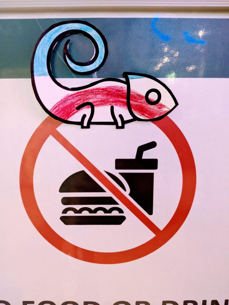 A no food or drink sign featuring an icon of a hamburger and drink which has been crossed out with a red circle and slash. A paper lizard has been colored to blend in with the top of the arc of the circle.